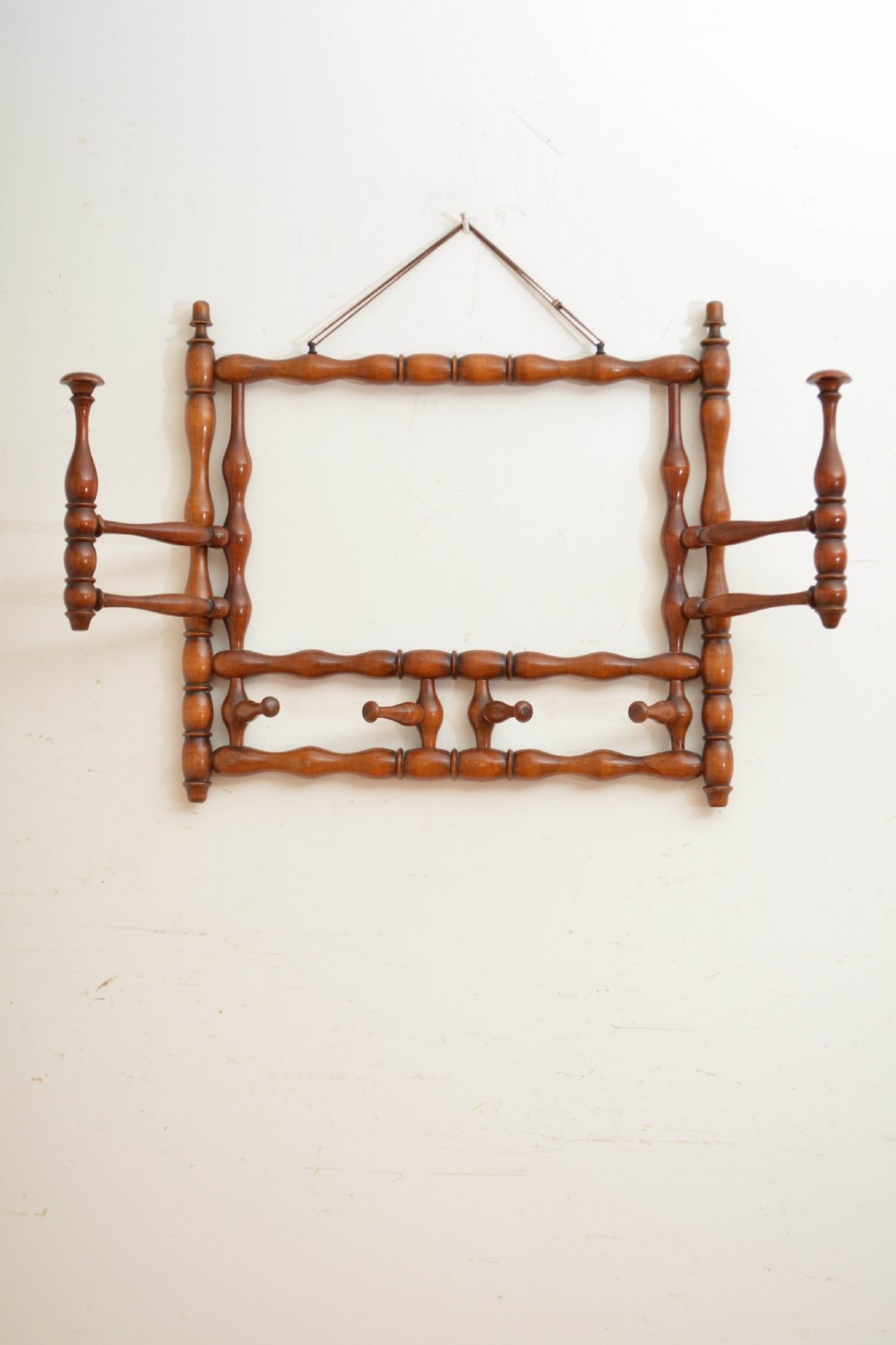 A wall-mounted coat rack made of turned warm brown wood spindles featuring multiple projecting hanging pegs and a top hanging cord. View II
