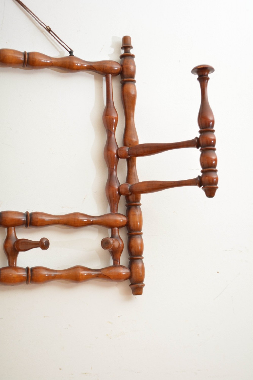 A wall-mounted coat rack made of turned warm brown wood spindles featuring multiple projecting hanging pegs and a top hanging cord. View III