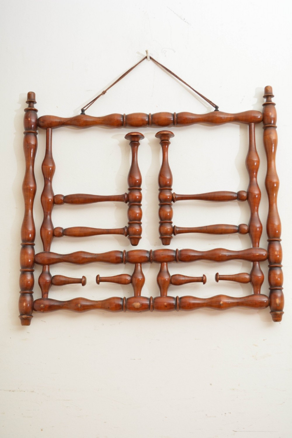 A wall-mounted coat rack made of turned warm brown wood spindles featuring multiple projecting hanging pegs and a top hanging cord. View V