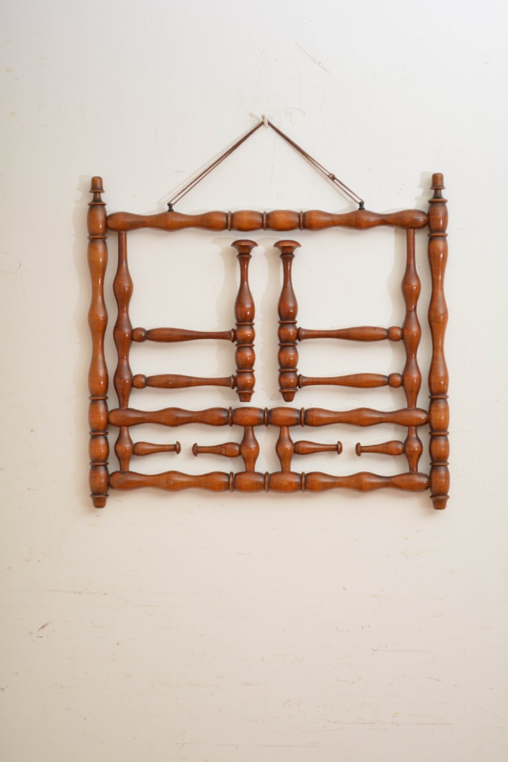 A wall-mounted coat rack made of turned warm brown wood spindles featuring multiple projecting hanging pegs and a top hanging cord.