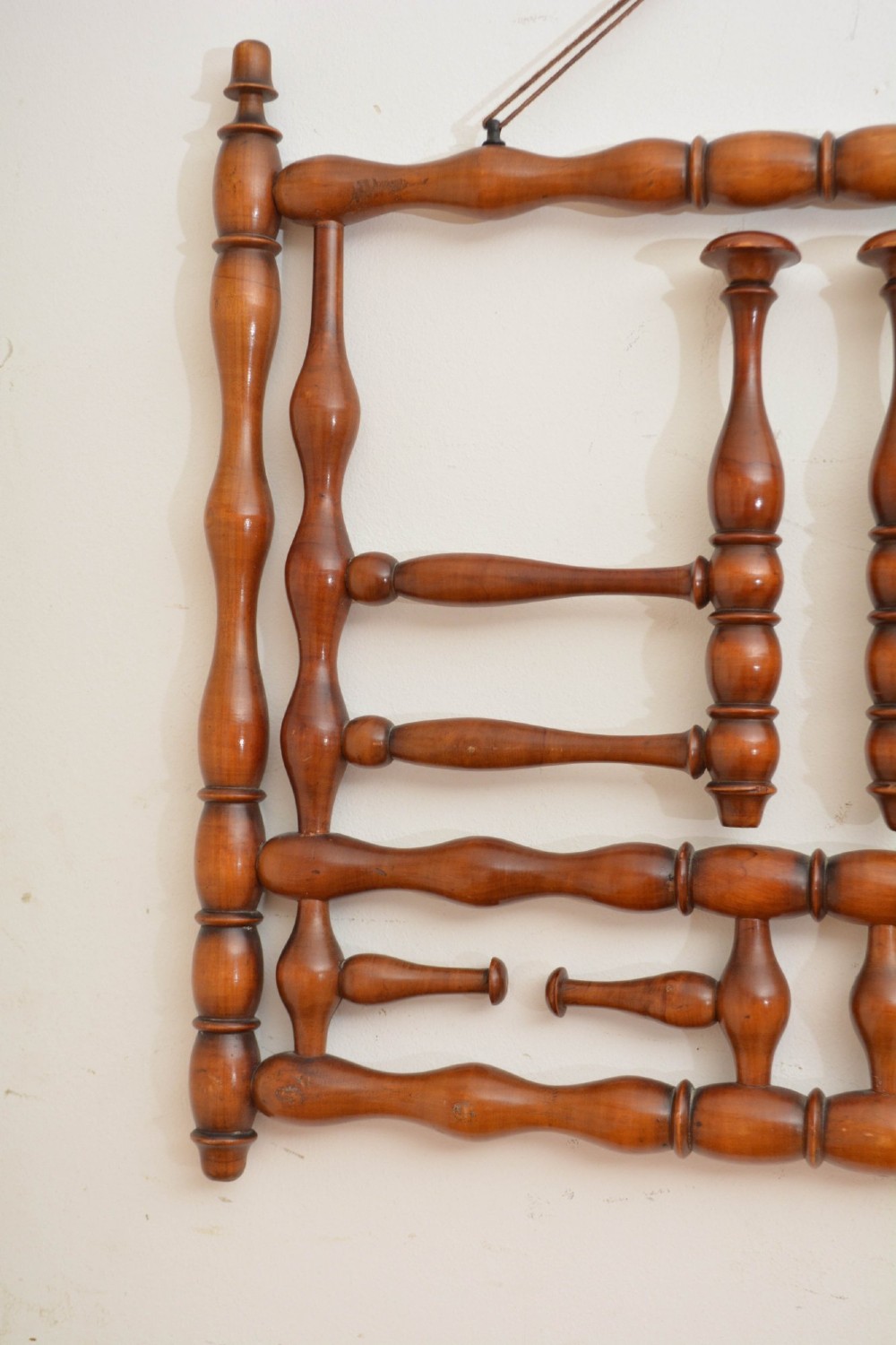 A wall-mounted coat rack made of turned warm brown wood spindles featuring multiple projecting hanging pegs and a top hanging cord. View VI