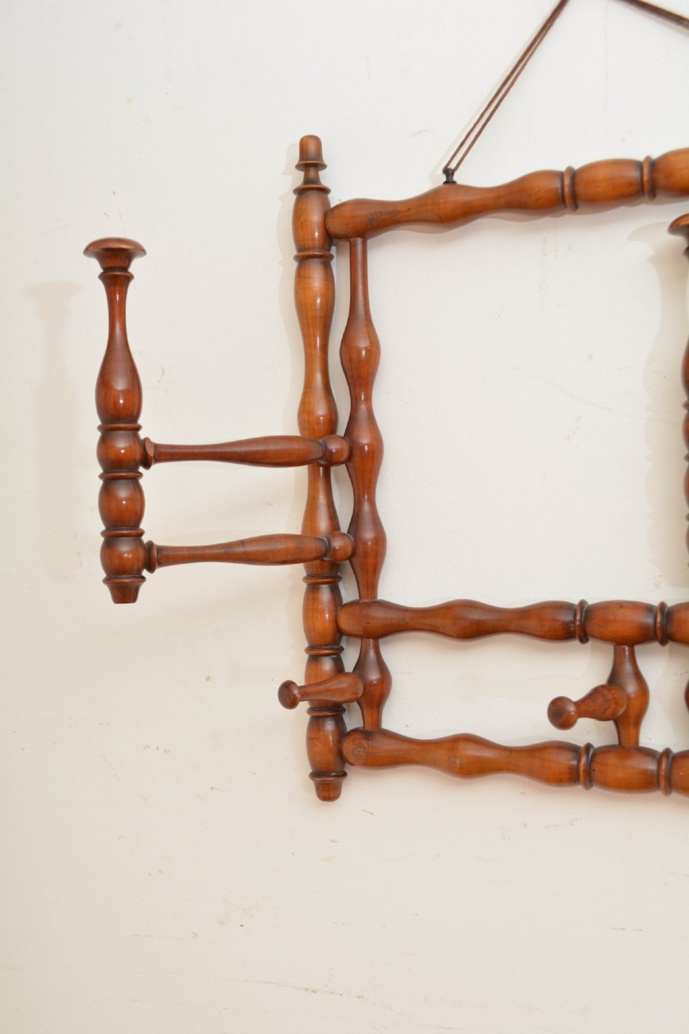 A wall-mounted coat rack made of turned warm brown wood spindles featuring multiple projecting hanging pegs and a top hanging cord. View VII