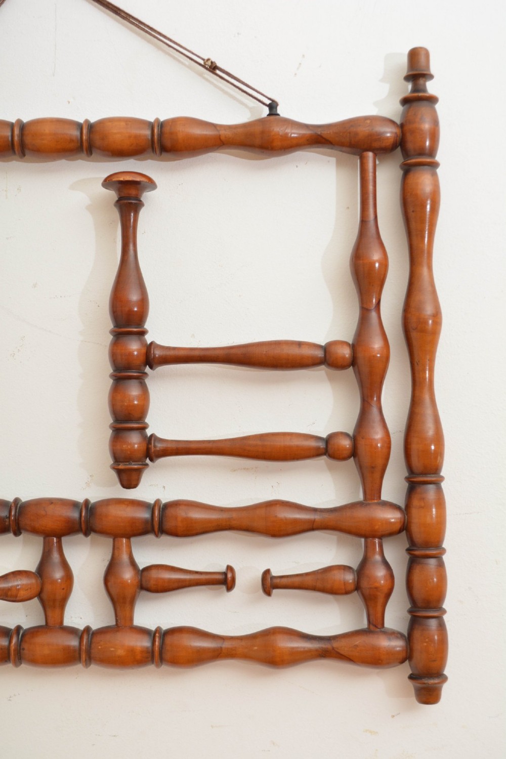 A wall-mounted coat rack made of turned warm brown wood spindles featuring multiple projecting hanging pegs and a top hanging cord. View X