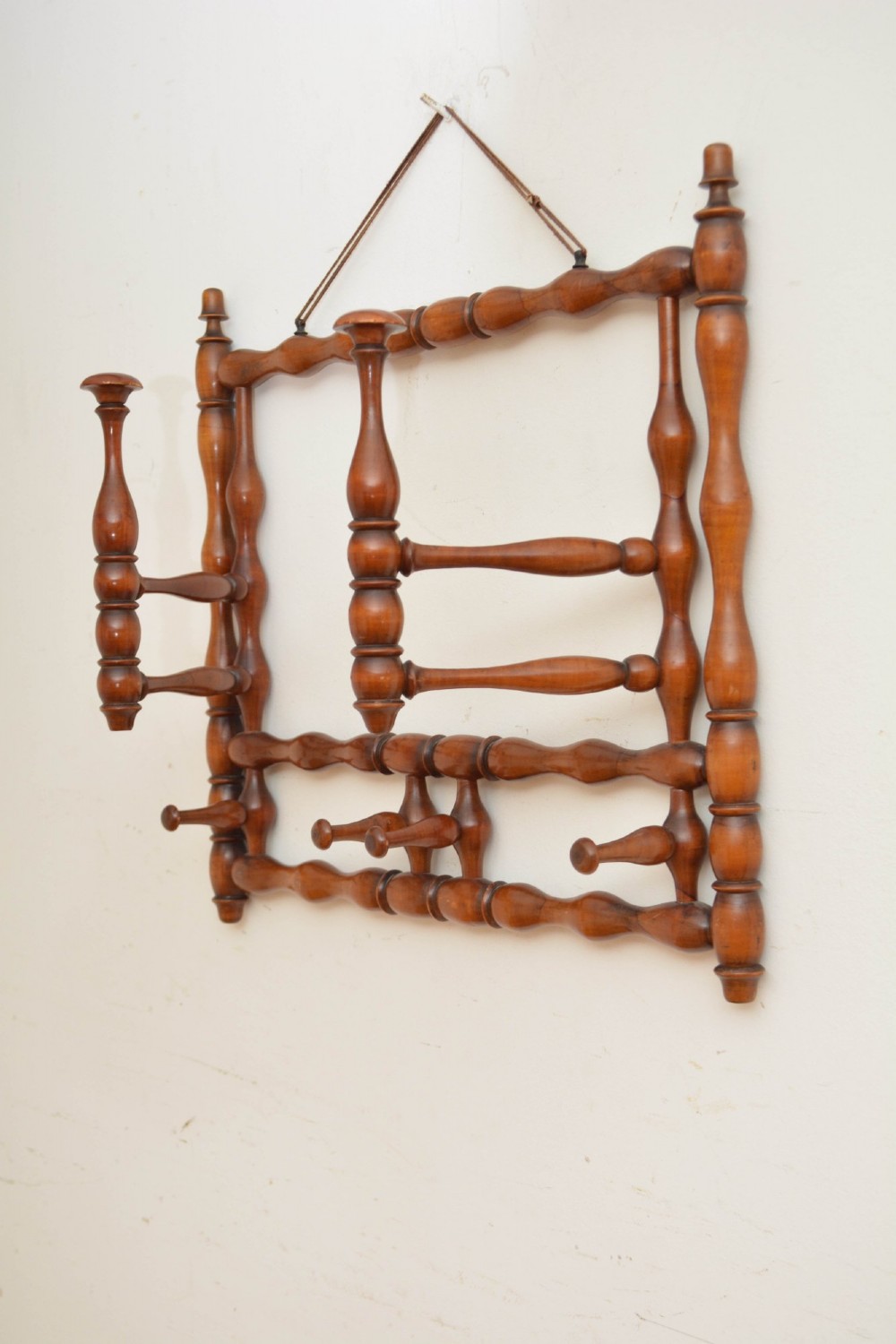 A wall-mounted coat rack made of turned warm brown wood spindles featuring multiple projecting hanging pegs and a top hanging cord. View XII