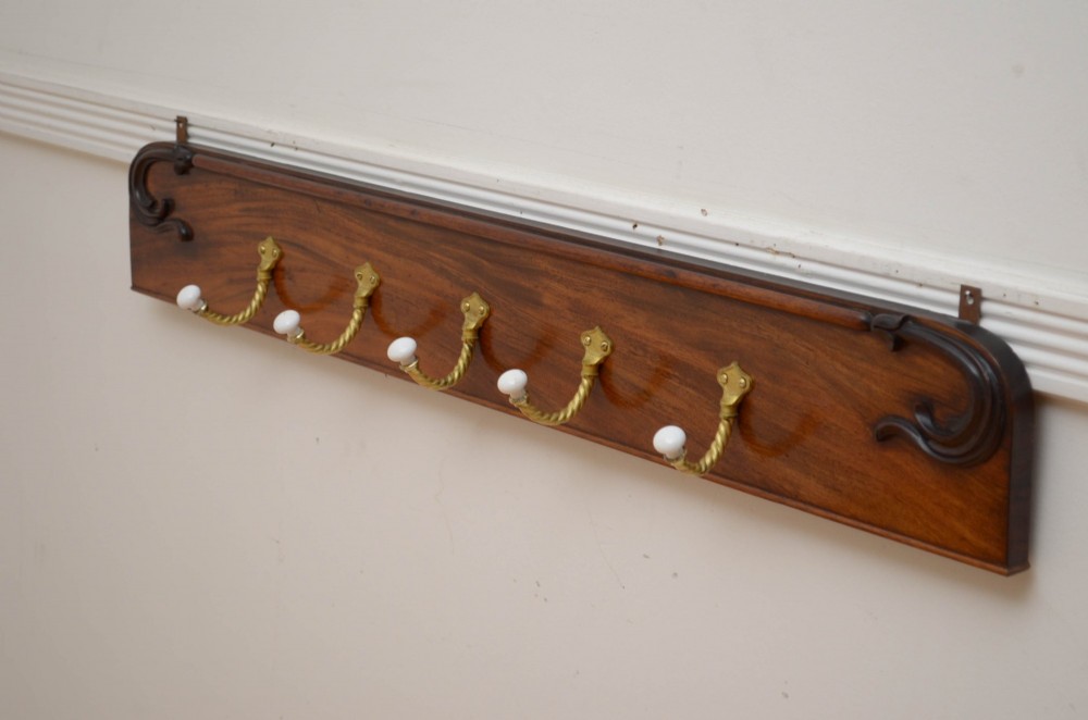An antique wall-mounted mahogany coat rack featuring five rope-twist brass hooks topped with white ceramic knobs and carved scrollwork details.