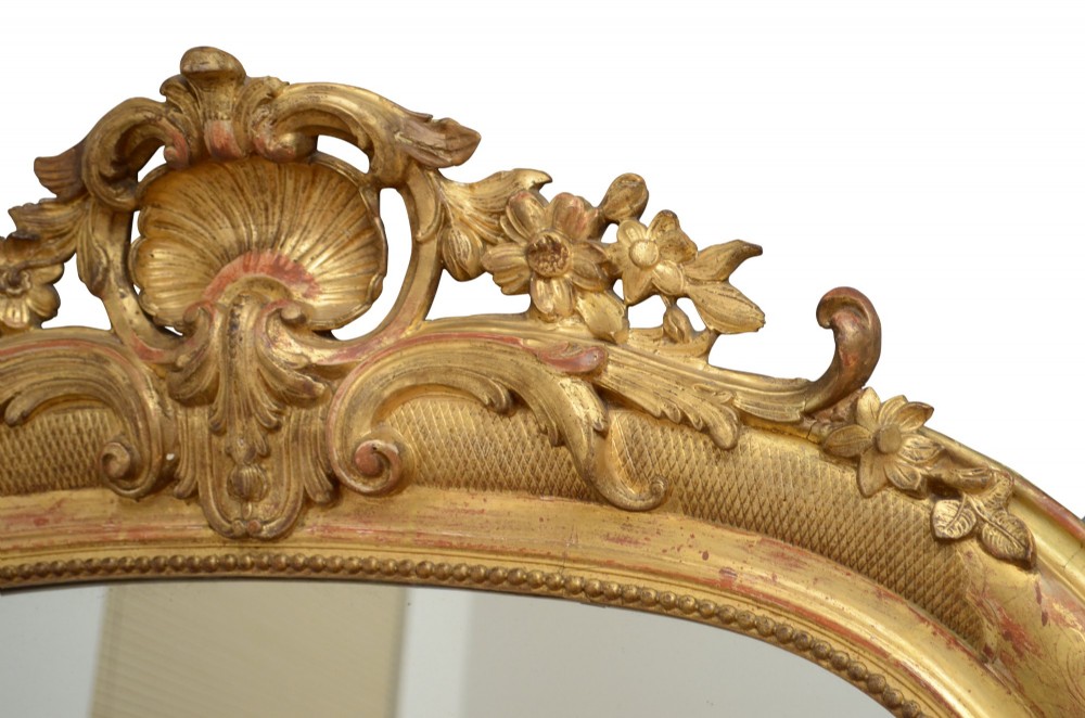 A tall arched giltwood wall mirror featuring an intricate shell cresting at the top and floral carved motifs along the gilded frame. View X