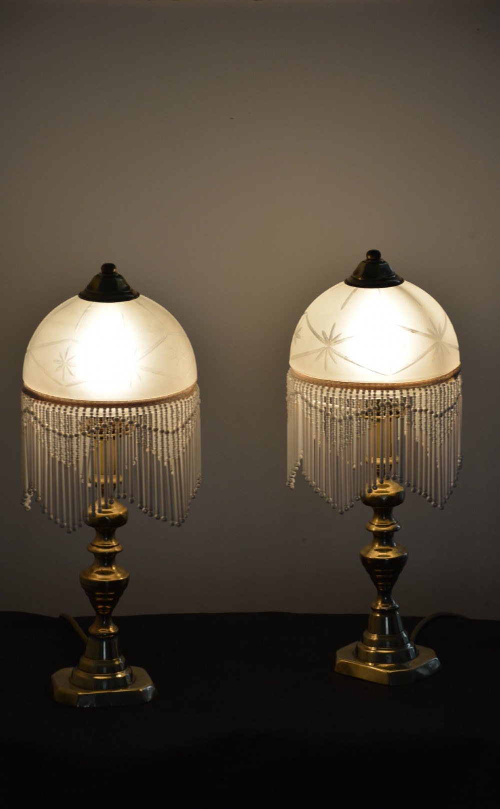A pair of illuminated Victorian turned brass table lamps featuring etched frosted glass dome shades with beaded fringe hanging around the rim.