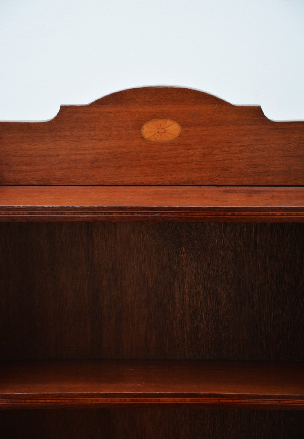 A slim Edwardian mahogany open bookcase featuring four shelves, an inlaid oval motif on the shaped gallery top, and bracket feet. View XV