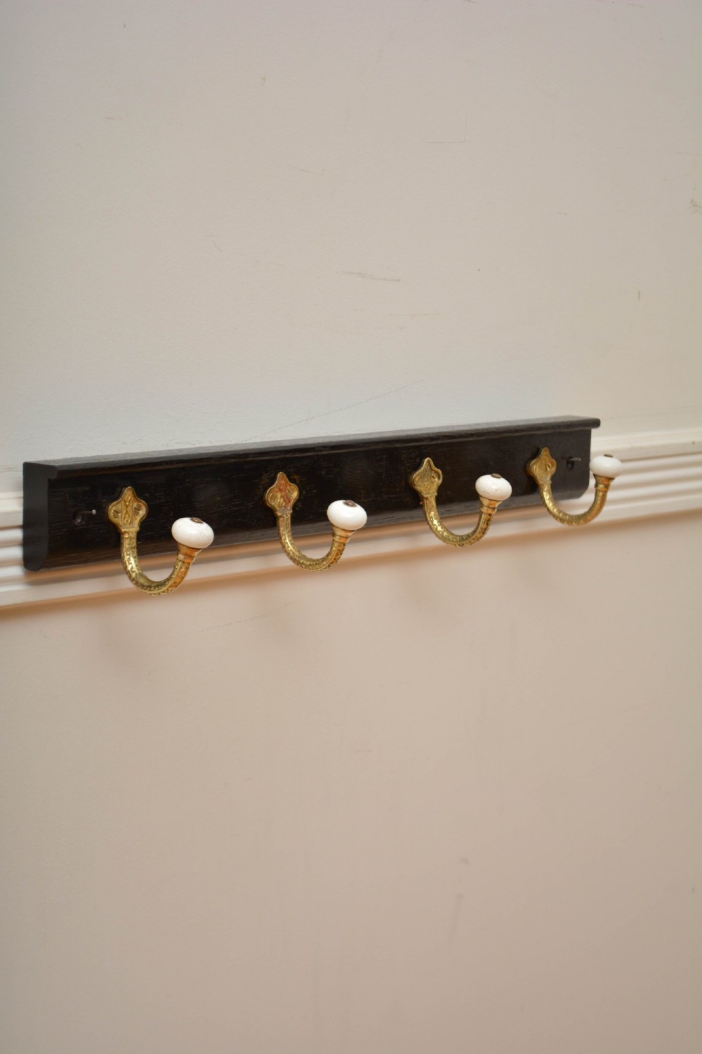 An ebonised black wooden wall rack featuring four cast brass hooks with white ceramic terminal knobs.