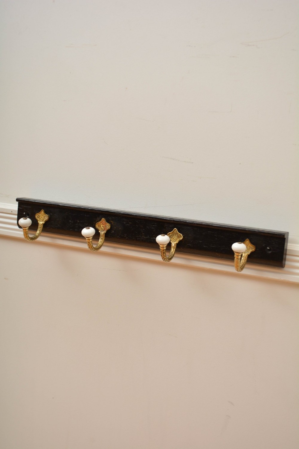 An ebonised black wooden wall rack featuring four cast brass hooks with white ceramic terminal knobs. View X