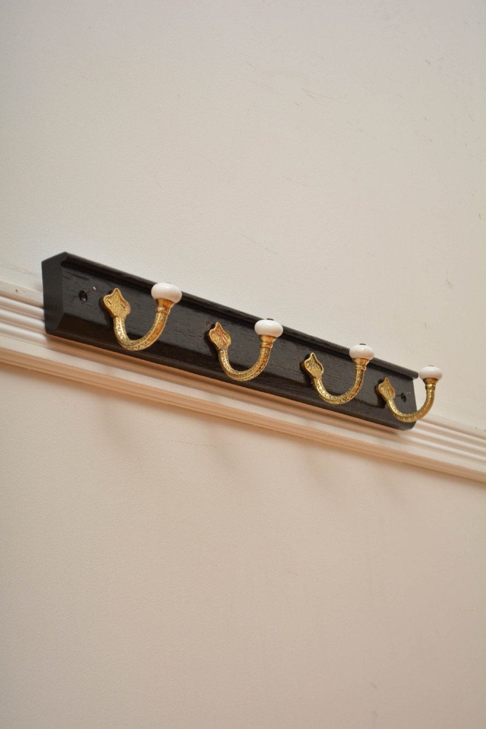 An ebonised black wooden wall rack featuring four cast brass hooks with white ceramic terminal knobs. View XI