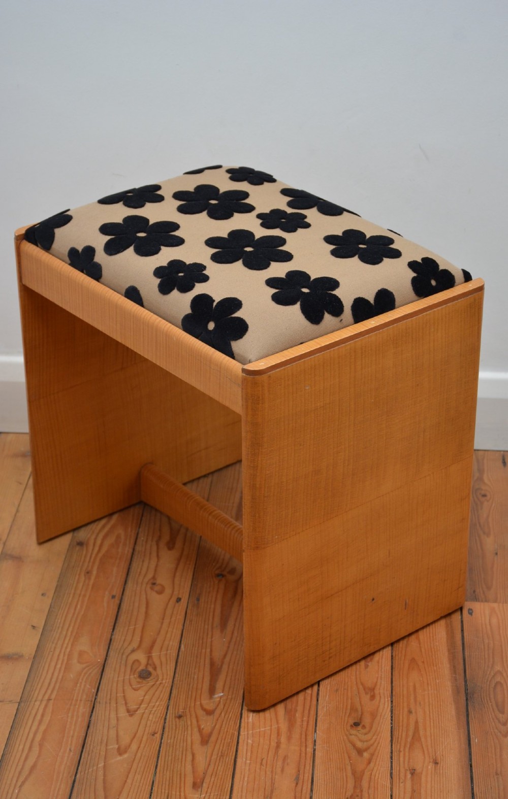 An Art Deco sycamore wood dressing stool featuring slab side legs, a low cylindrical stretcher, and a padded seat upholstered in beige fabric with black floral motifs. View II