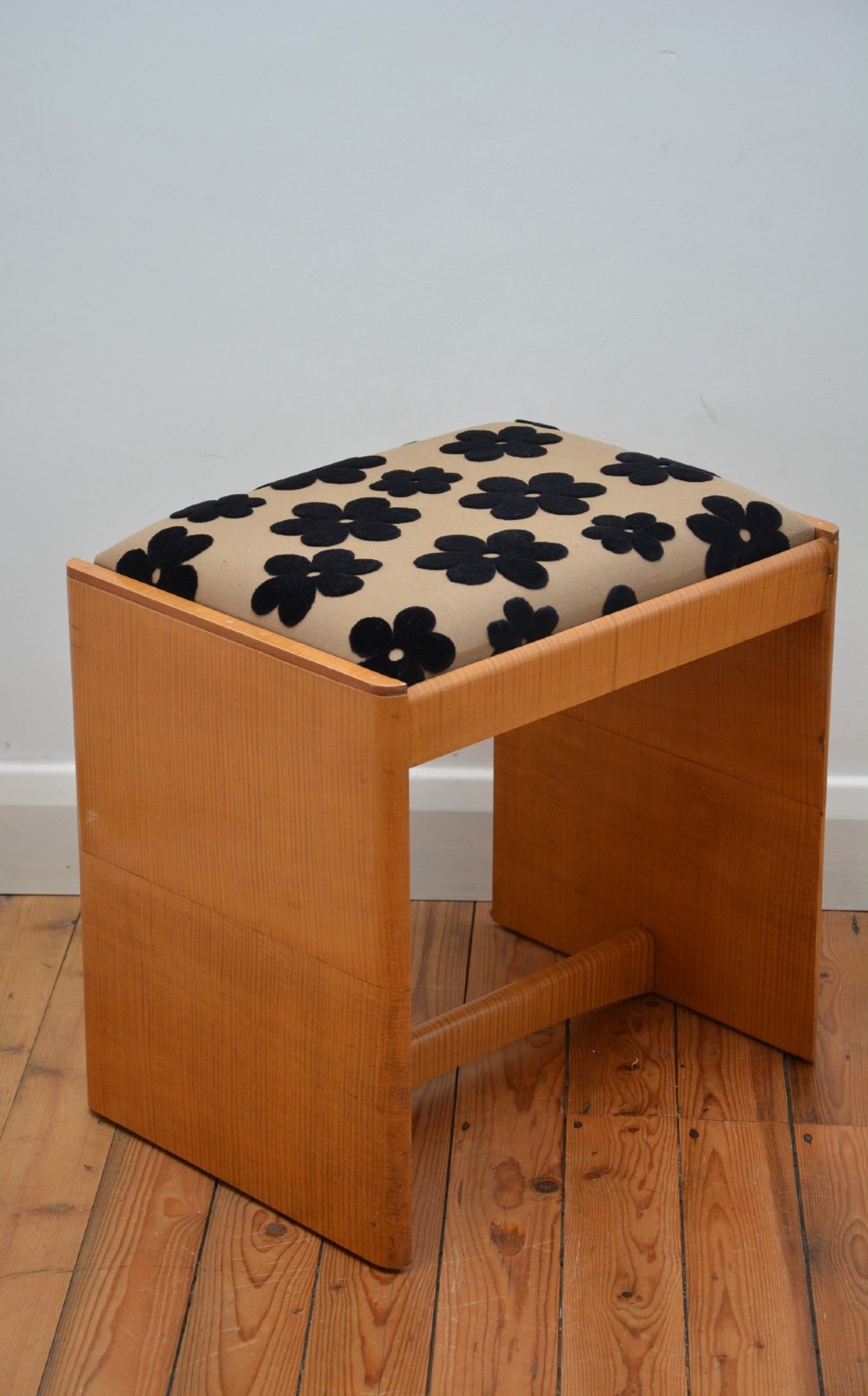 An Art Deco sycamore wood dressing stool featuring slab side legs, a low cylindrical stretcher, and a padded seat upholstered in beige fabric with black floral motifs.