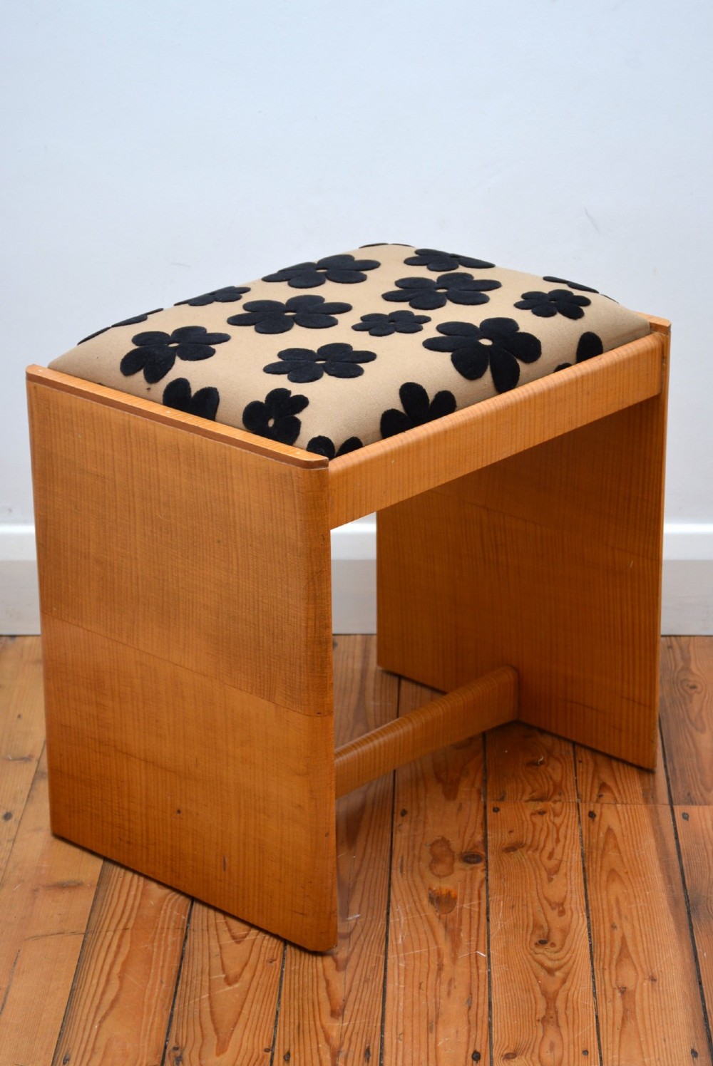 An Art Deco sycamore wood dressing stool featuring slab side legs, a low cylindrical stretcher, and a padded seat upholstered in beige fabric with black floral motifs. View X