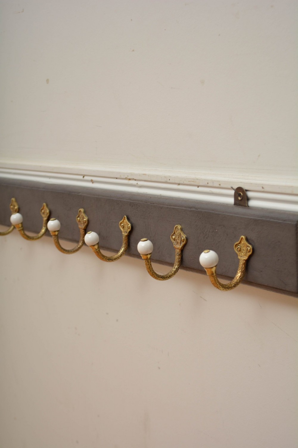 A long dark timber wall-mounted coat rack featuring multiple cast brass hooks with white ceramic finials. View VI