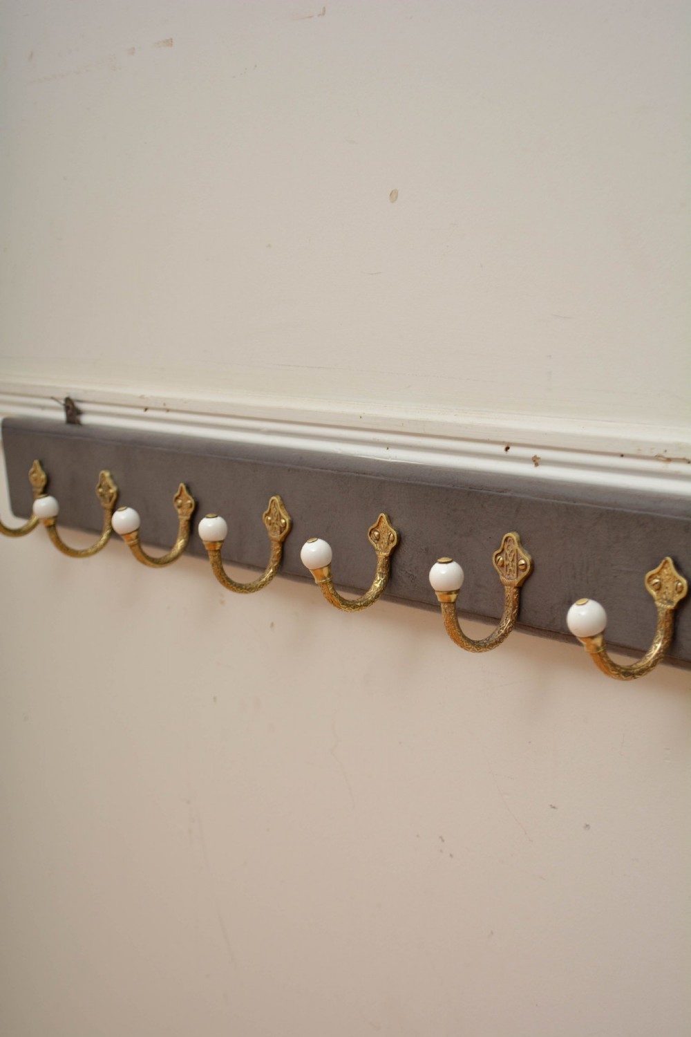 A long dark timber wall-mounted coat rack featuring multiple cast brass hooks with white ceramic finials. View VII
