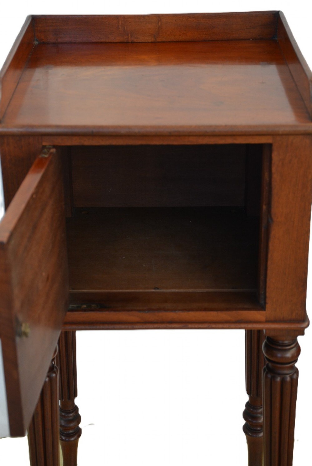 A dark mahogany bedside cabinet featuring a three-sided gallery top, a single door with a turned wooden knob, and four slender reeded legs. View X
