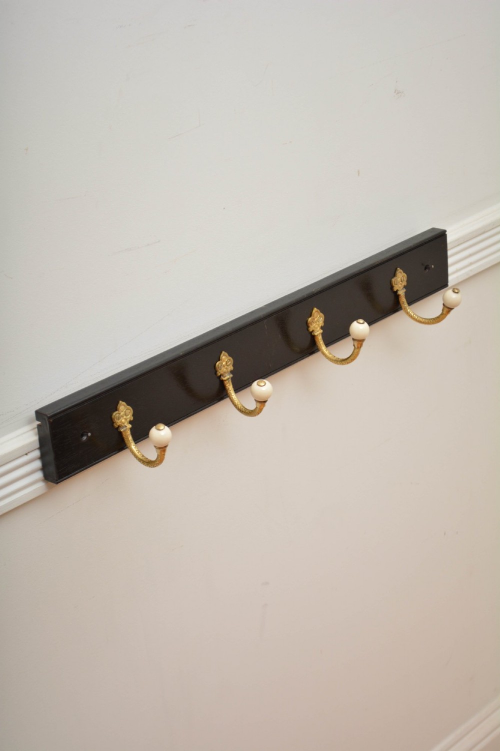 An ebonised black timber rail mounted with four cast brass coat hooks topped with round cream ceramic tips. View X