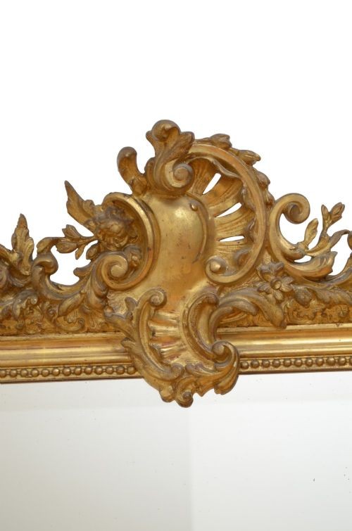 A tall antique giltwood wall mirror featuring an ornate carved floral cresting at the top, decorative foliate moulding along the frame, and original glass. View V