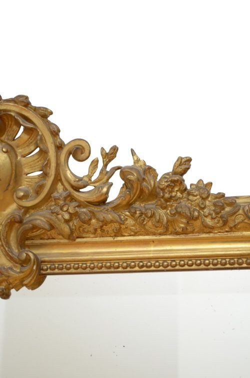 A tall antique giltwood wall mirror featuring an ornate carved floral cresting at the top, decorative foliate moulding along the frame, and original glass. View VI