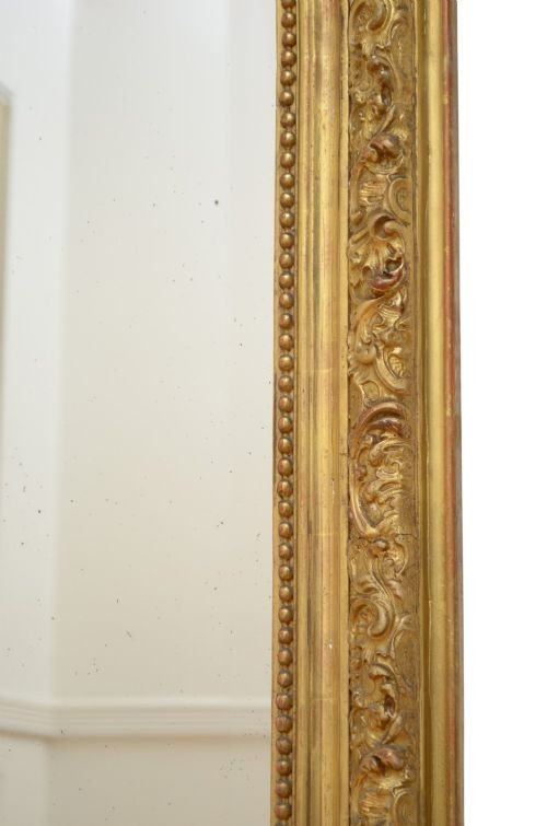 A tall antique giltwood wall mirror featuring an ornate carved floral cresting at the top, decorative foliate moulding along the frame, and original glass. View IX