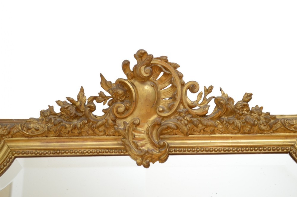 A tall antique giltwood wall mirror featuring an ornate carved floral cresting at the top, decorative foliate moulding along the frame, and original glass. View X