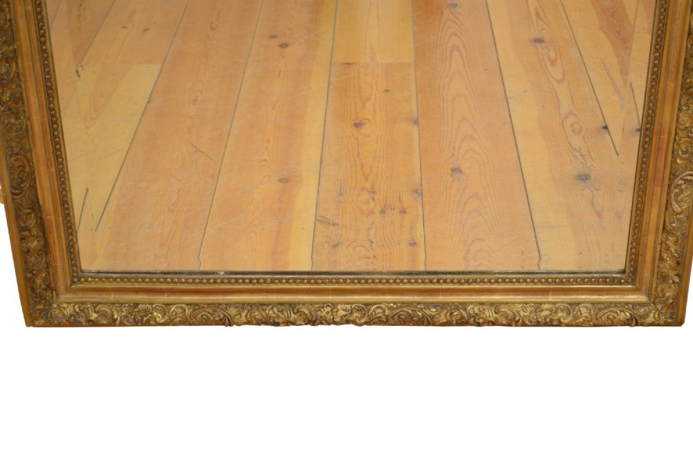 A tall antique giltwood wall mirror featuring an ornate carved floral cresting at the top, decorative foliate moulding along the frame, and original glass. View XI