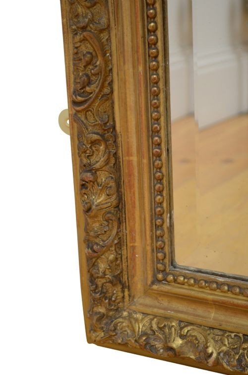 A tall antique giltwood wall mirror featuring an ornate carved floral cresting at the top, decorative foliate moulding along the frame, and original glass. View XV