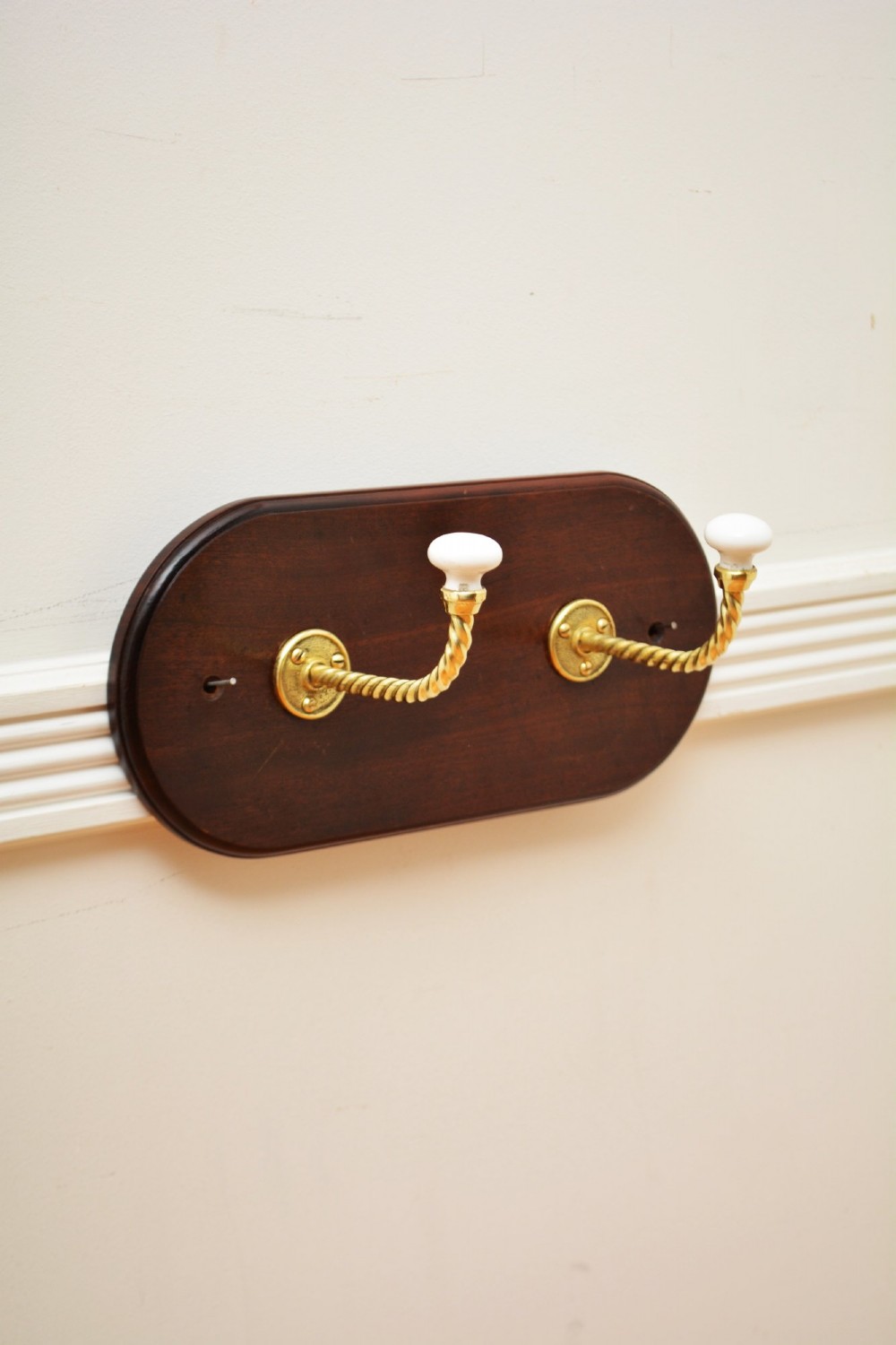 An oval mahogany wood backboard mounted with two twisted brass coat hooks featuring white ceramic end knobs.
