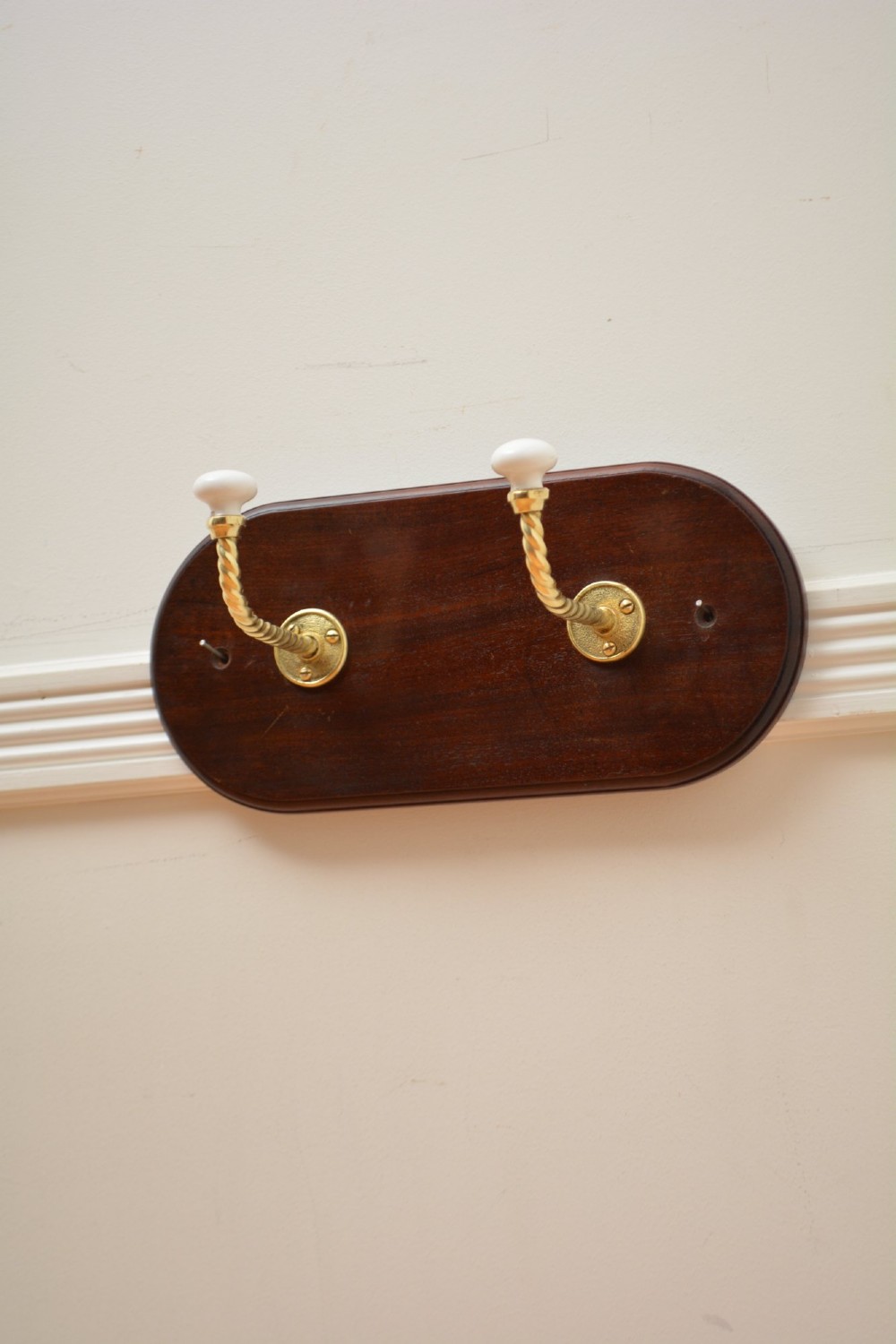 An oval mahogany wood backboard mounted with two twisted brass coat hooks featuring white ceramic end knobs. View IV