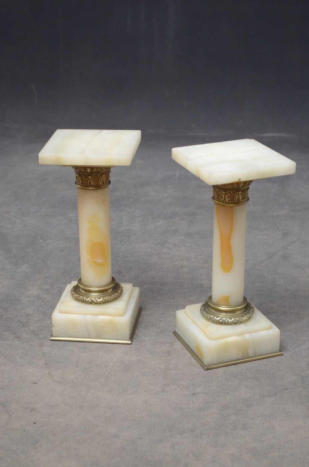 A pair of low Neoclassical onyx pedestal columns with square stepped bases, cylindrical stone shafts, and decorative brass capitals. View II