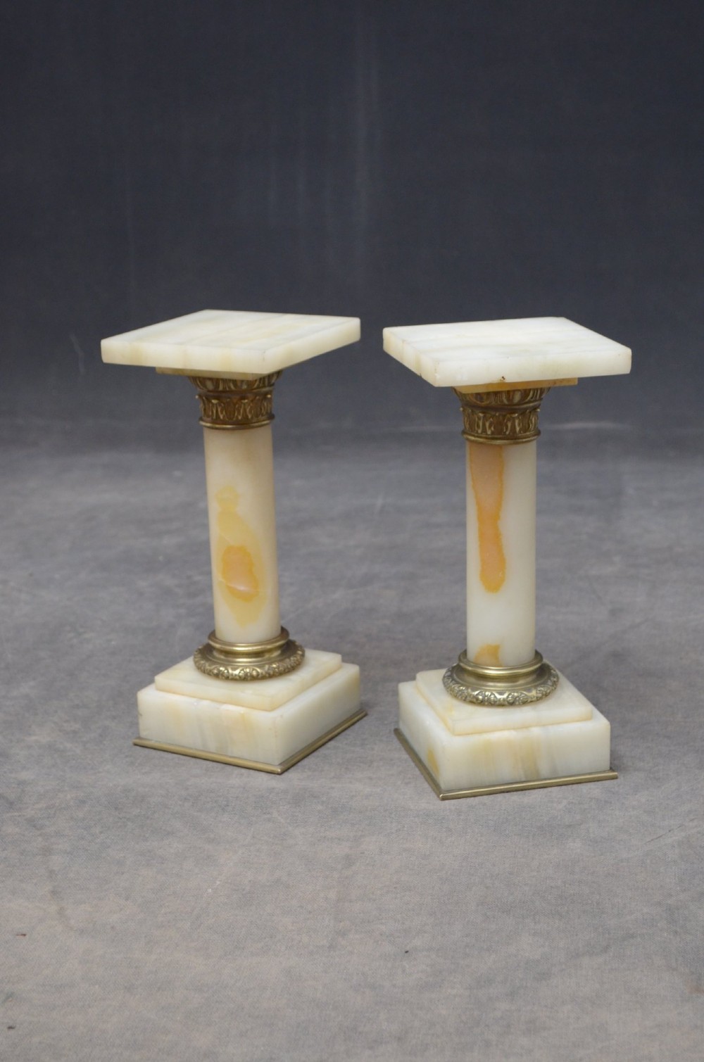 A pair of low Neoclassical onyx pedestal columns with square stepped bases, cylindrical stone shafts, and decorative brass capitals. View III