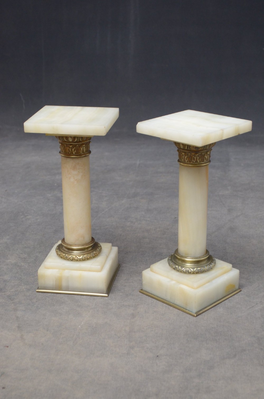 A pair of low Neoclassical onyx pedestal columns with square stepped bases, cylindrical stone shafts, and decorative brass capitals. View IV