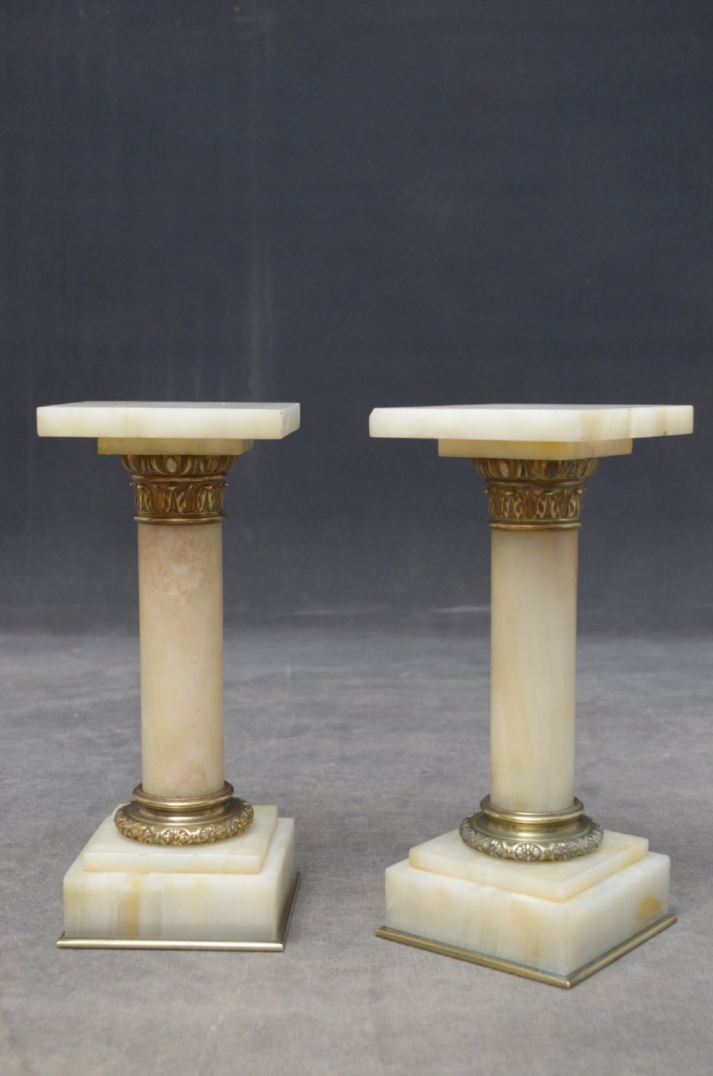 A pair of low Neoclassical onyx pedestal columns with square stepped bases, cylindrical stone shafts, and decorative brass capitals. View V