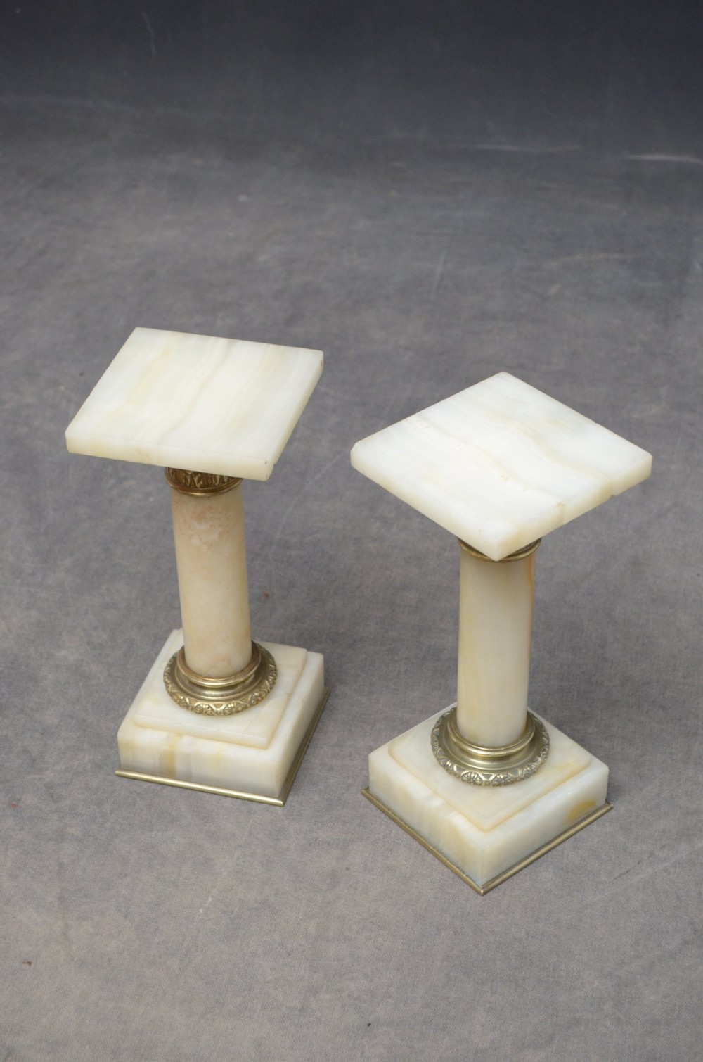 A pair of low Neoclassical onyx pedestal columns with square stepped bases, cylindrical stone shafts, and decorative brass capitals. View VI