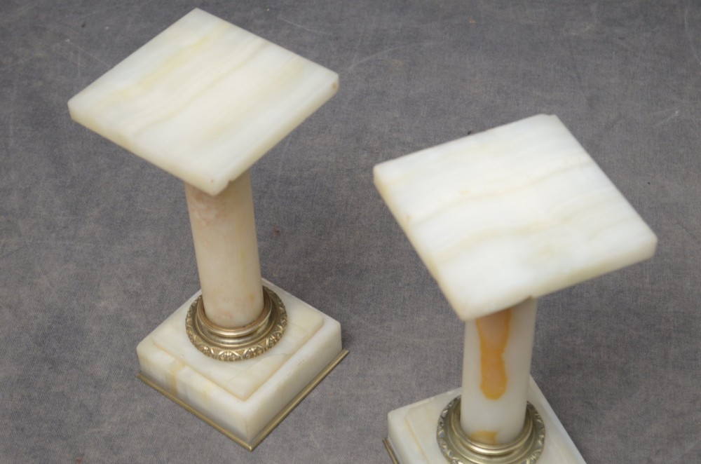 A pair of low Neoclassical onyx pedestal columns with square stepped bases, cylindrical stone shafts, and decorative brass capitals. View VIII