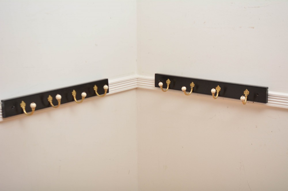 A pair of wall-mounted ebonised black wooden rails fitted with curved brass coat hooks ending in white ceramic knobs.