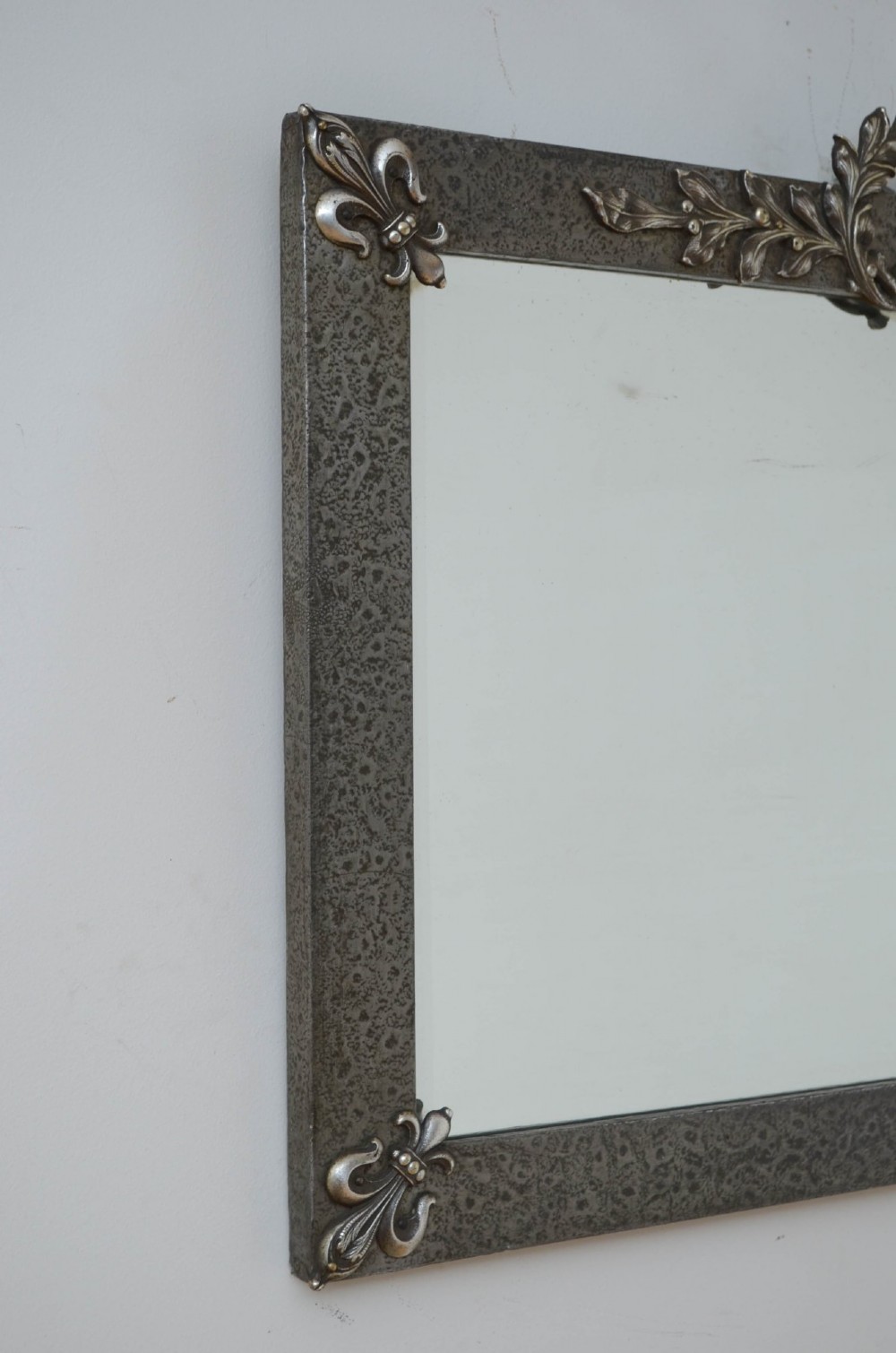 A rectangular Arts and Crafts wall mirror with a hand-hammered silver metal frame, decorated with fleur-de-lis corner accents and a foliate wreath crest. View II