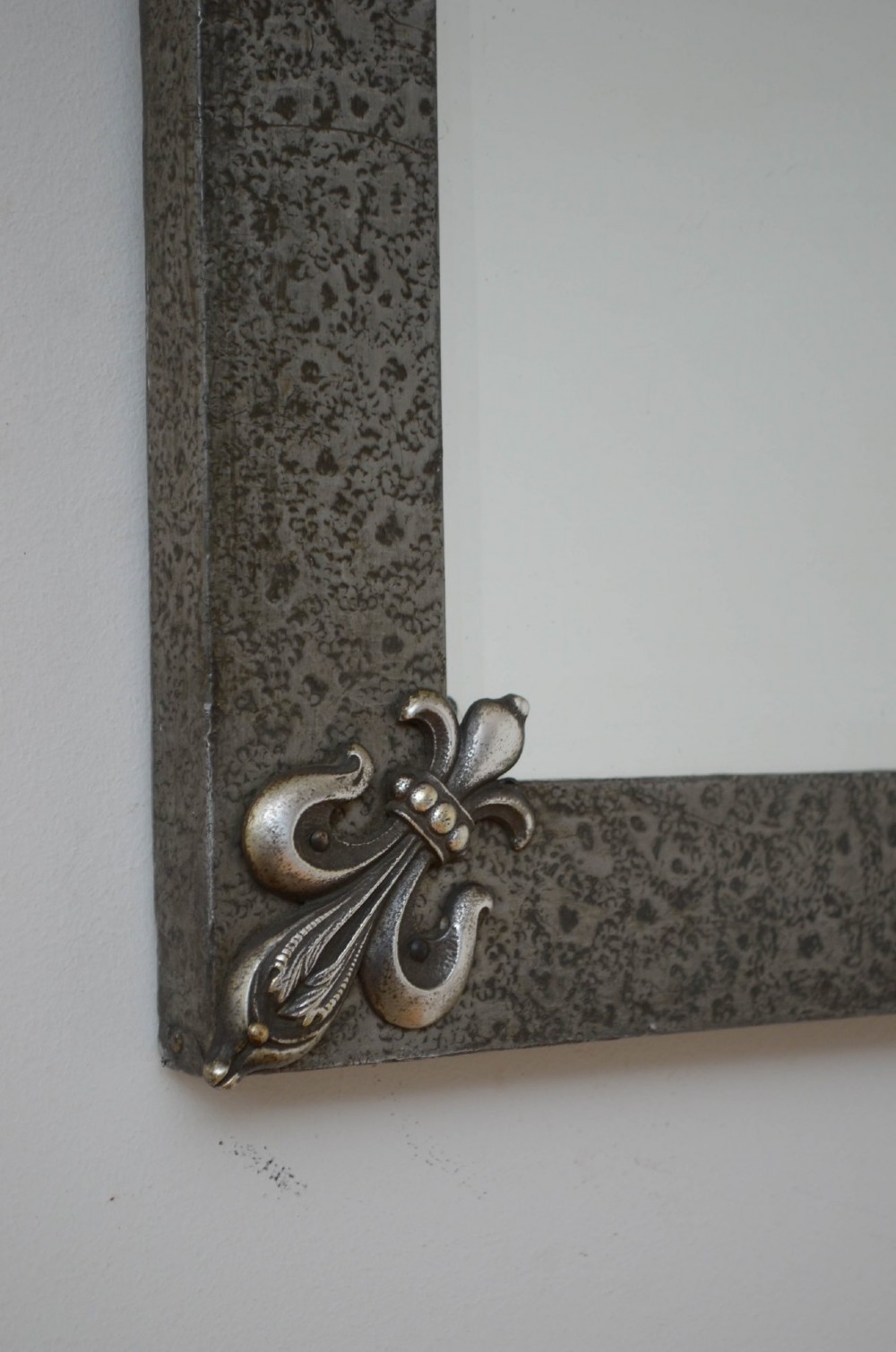 A rectangular Arts and Crafts wall mirror with a hand-hammered silver metal frame, decorated with fleur-de-lis corner accents and a foliate wreath crest. View III