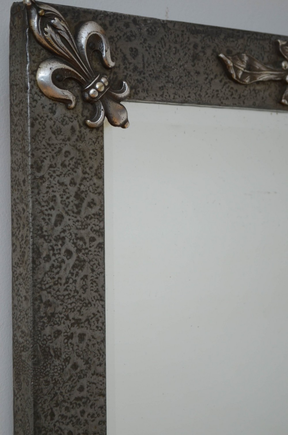 A rectangular Arts and Crafts wall mirror with a hand-hammered silver metal frame, decorated with fleur-de-lis corner accents and a foliate wreath crest. View IV