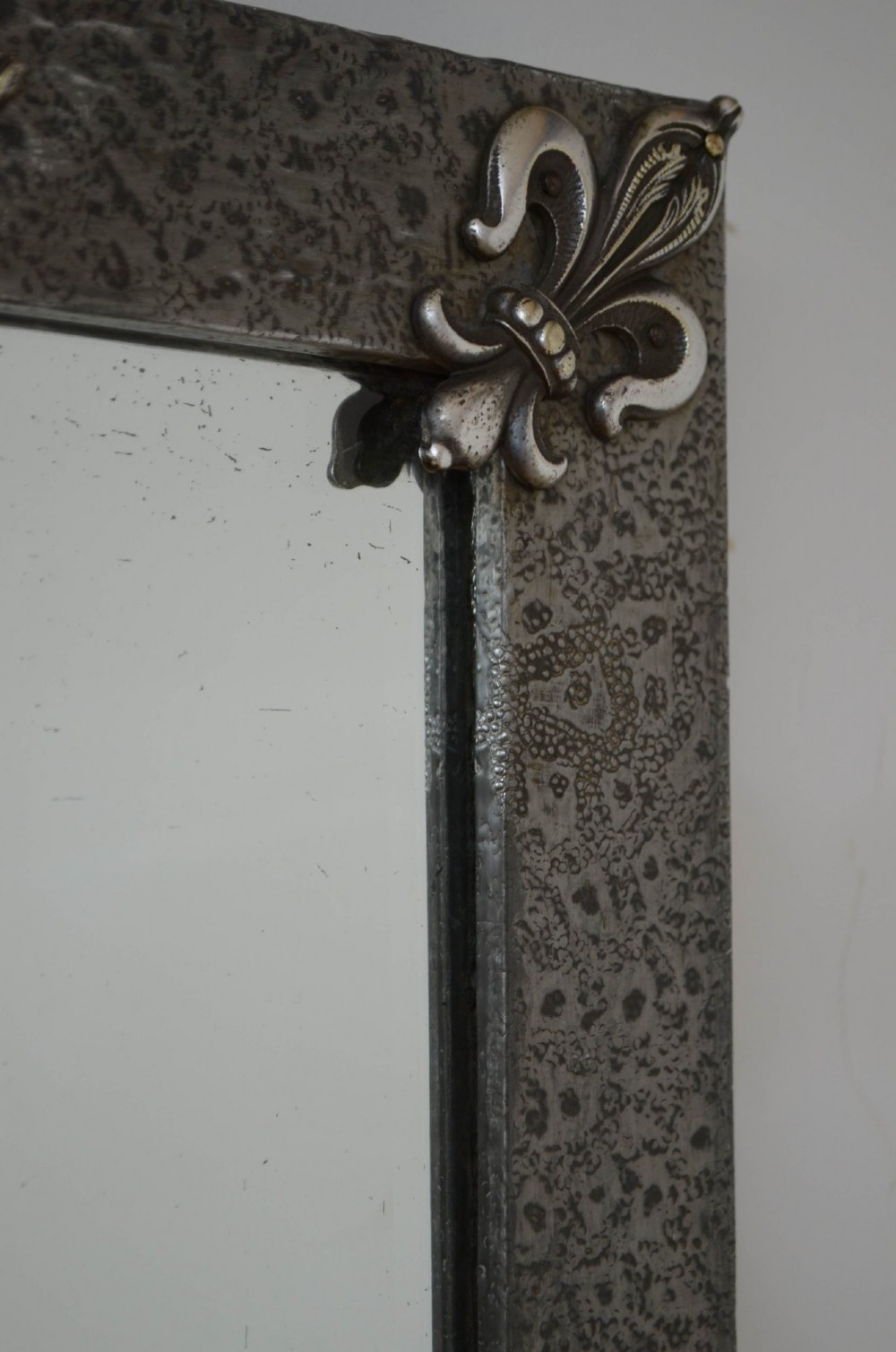 A rectangular Arts and Crafts wall mirror with a hand-hammered silver metal frame, decorated with fleur-de-lis corner accents and a foliate wreath crest. View VI