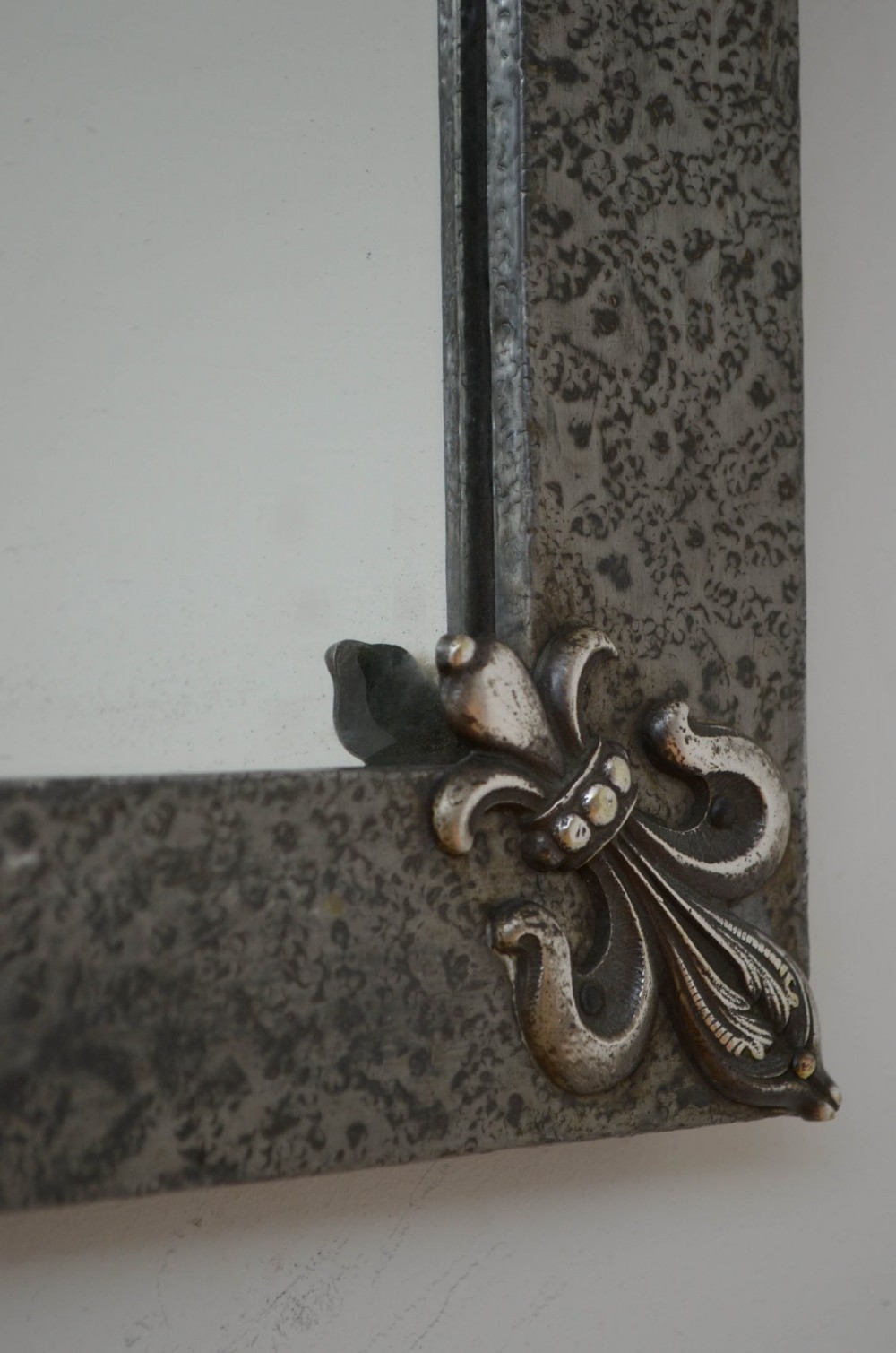 A rectangular Arts and Crafts wall mirror with a hand-hammered silver metal frame, decorated with fleur-de-lis corner accents and a foliate wreath crest. View VII
