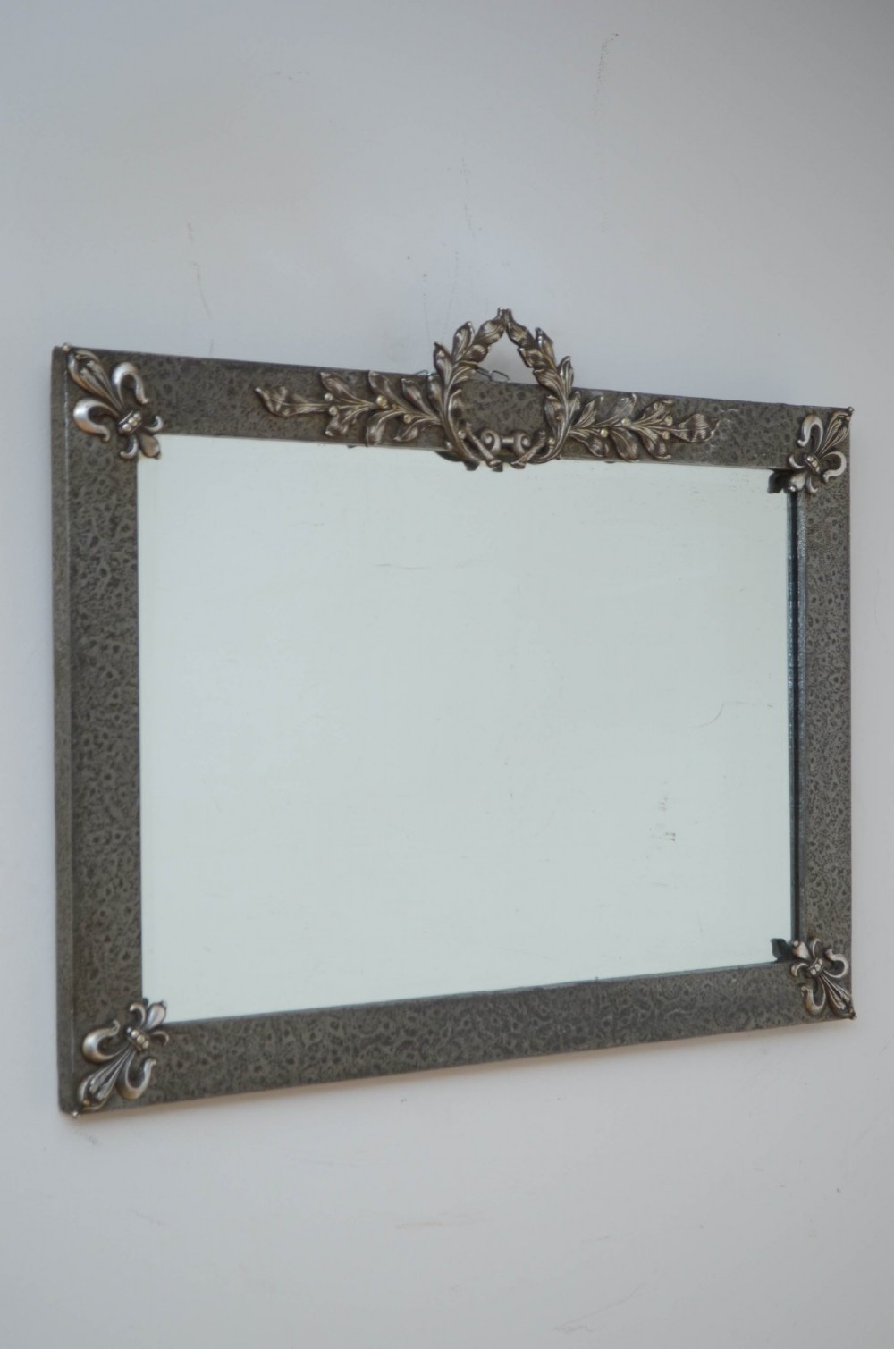 A rectangular Arts and Crafts wall mirror with a hand-hammered silver metal frame, decorated with fleur-de-lis corner accents and a foliate wreath crest. View IX