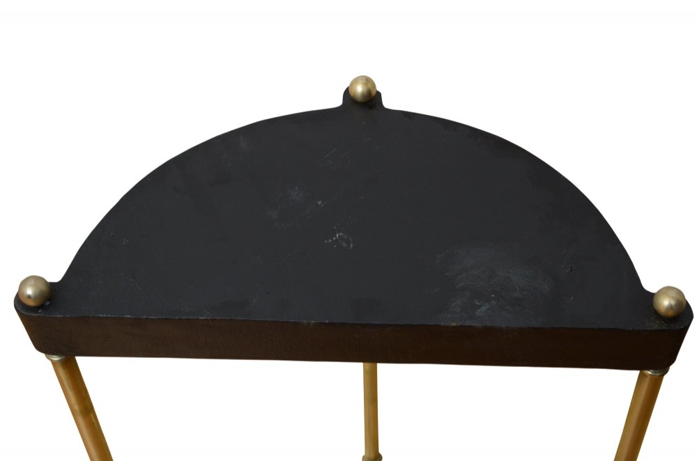 A semi-circular brass umbrella stand featuring vertical tubular supports, decorative finials, top divisions, and a black cast-iron drip tray base. View VII