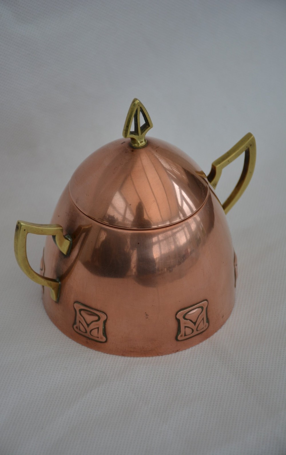 An Art Nouveau copper sugar bowl featuring angular brass handles, a conical lid with a stylised finial, and embossed geometric motifs around the base. View II