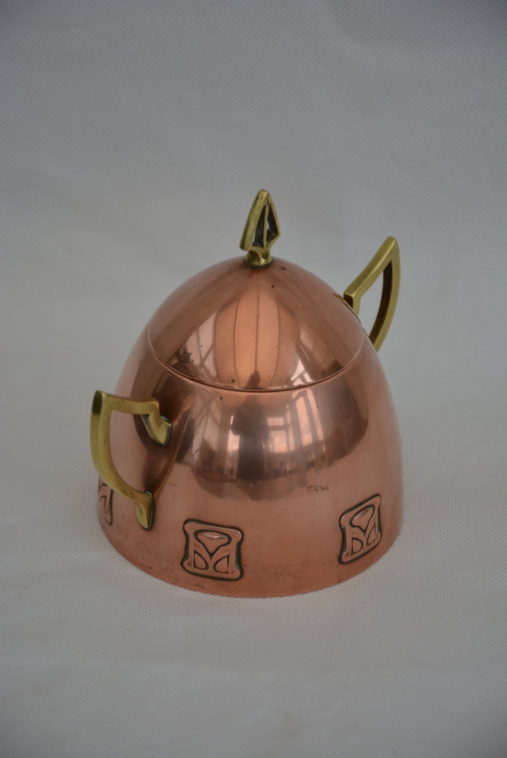 An Art Nouveau copper sugar bowl featuring angular brass handles, a conical lid with a stylised finial, and embossed geometric motifs around the base. View V
