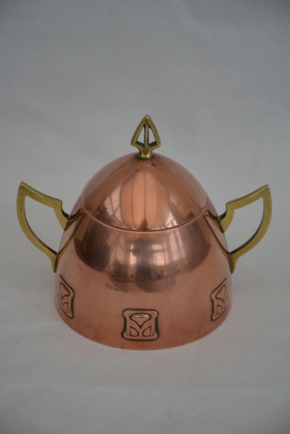 An Art Nouveau copper sugar bowl featuring angular brass handles, a conical lid with a stylised finial, and embossed geometric motifs around the base. View VI