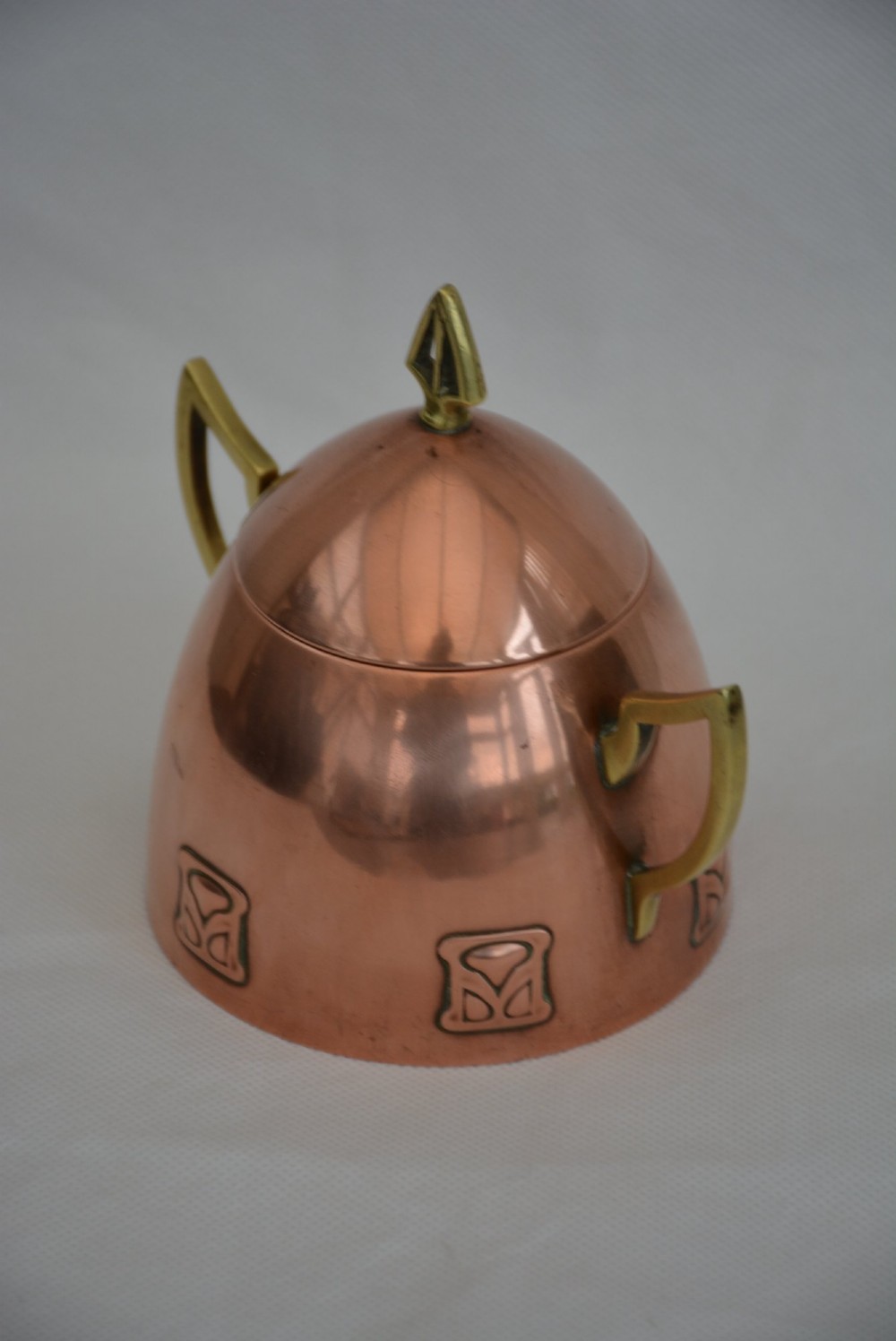 An Art Nouveau copper sugar bowl featuring angular brass handles, a conical lid with a stylised finial, and embossed geometric motifs around the base. View VII