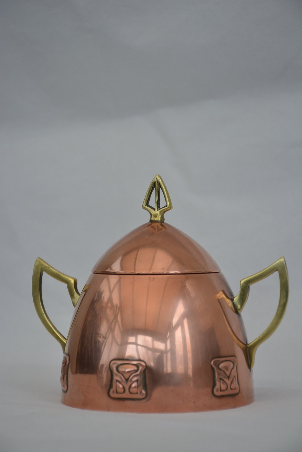 An Art Nouveau copper sugar bowl featuring angular brass handles, a conical lid with a stylised finial, and embossed geometric motifs around the base. View VIII