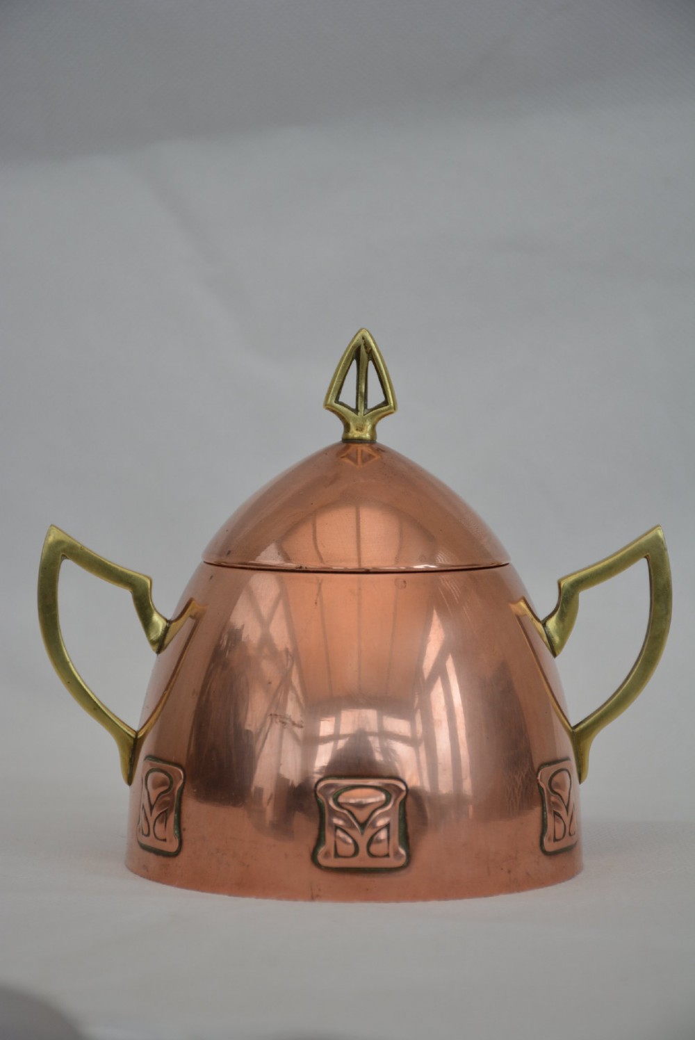 An Art Nouveau copper sugar bowl featuring angular brass handles, a conical lid with a stylised finial, and embossed geometric motifs around the base. View IX